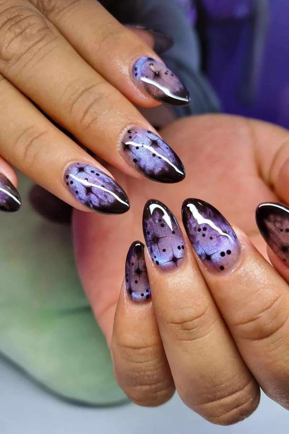 Close-up of hands with almond-shaped nails painted in a purple and black marbled design with shiny finish and small black dot accents.