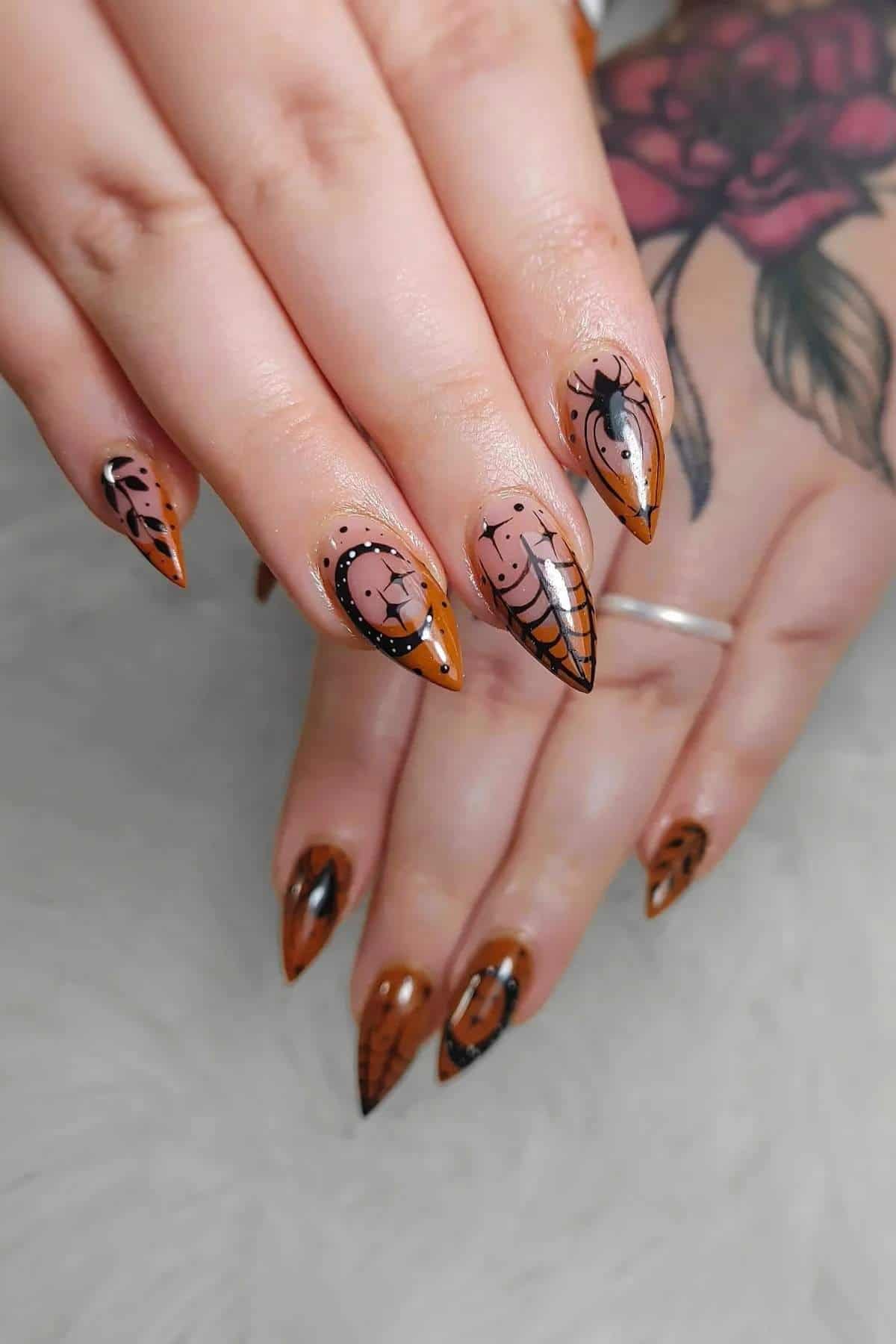 Hand with long, pointed nails featuring Halloween-themed nail art, including spider webs, spiders, and moons, in black and orange tones. Tattoo visible on hand and wrist.