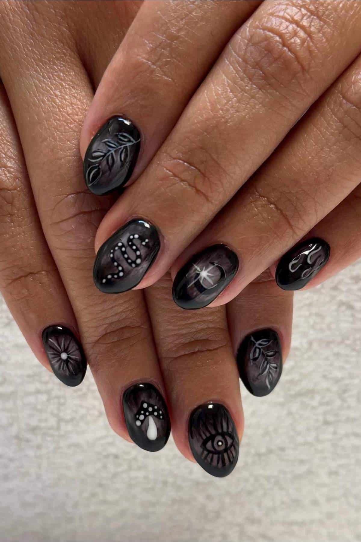 Close-up of hands with black nails featuring white and gray designs, including an eye, leaves, a heart, a daisy, and abstract patterns.