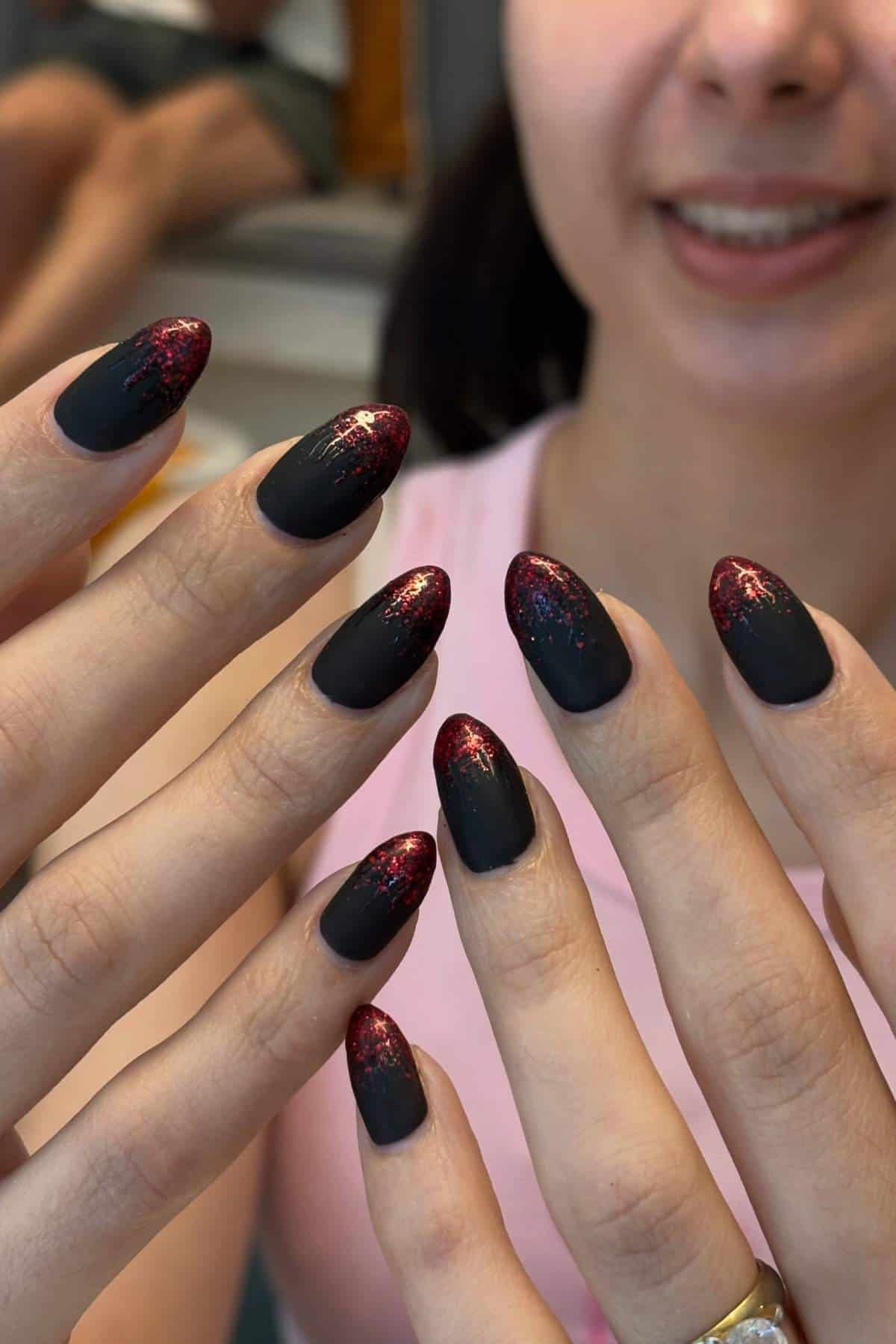 Woman showing hands with almond-shaped nails painted matte black with red glitter tips. She is smiling, wearing a pink top, and has a gold ring on her finger.
