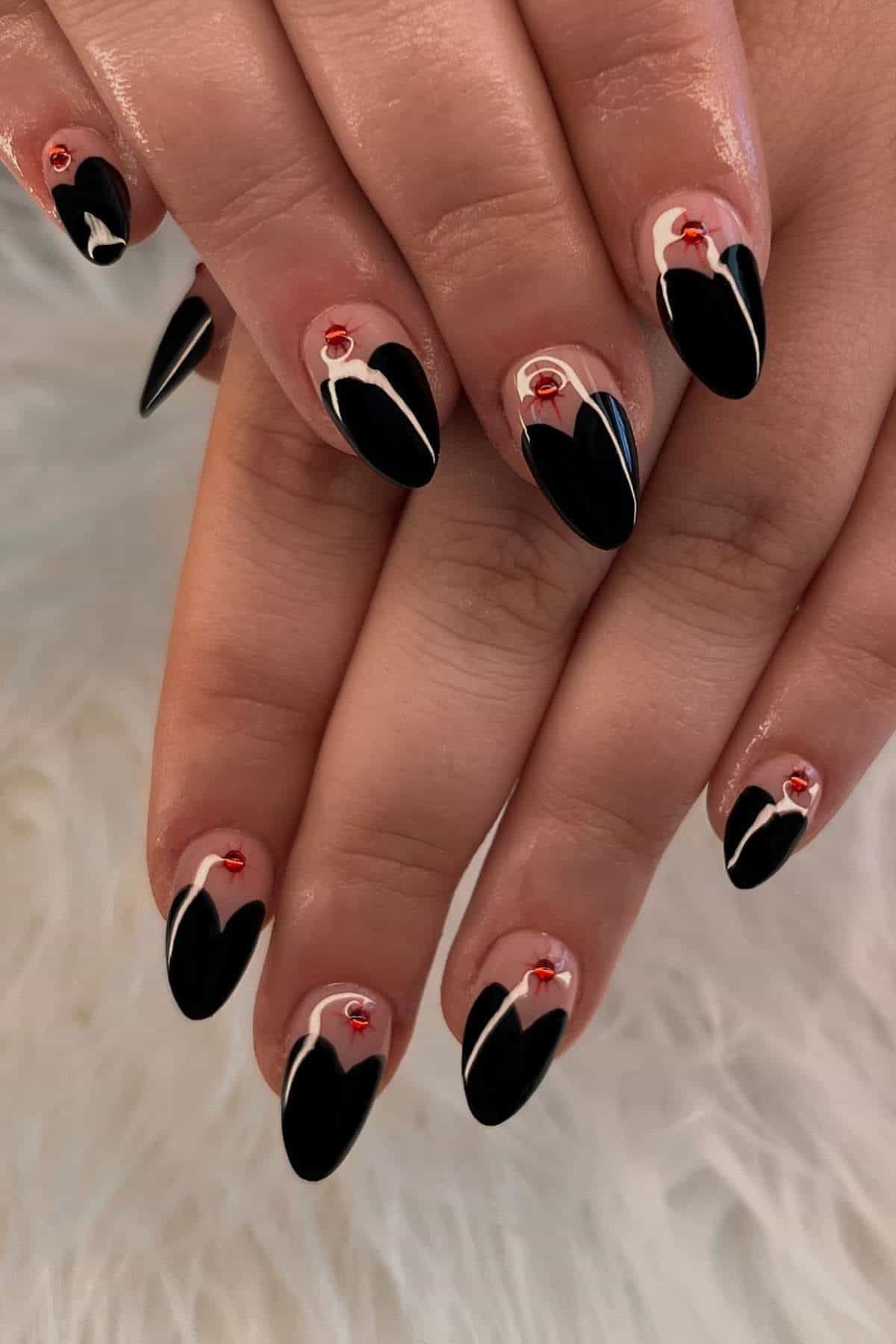 Hands with almond-shaped nails featuring black heart tips, white outlines, and small red rhinestone accents on a nude background, resting on a fluffy white surface.