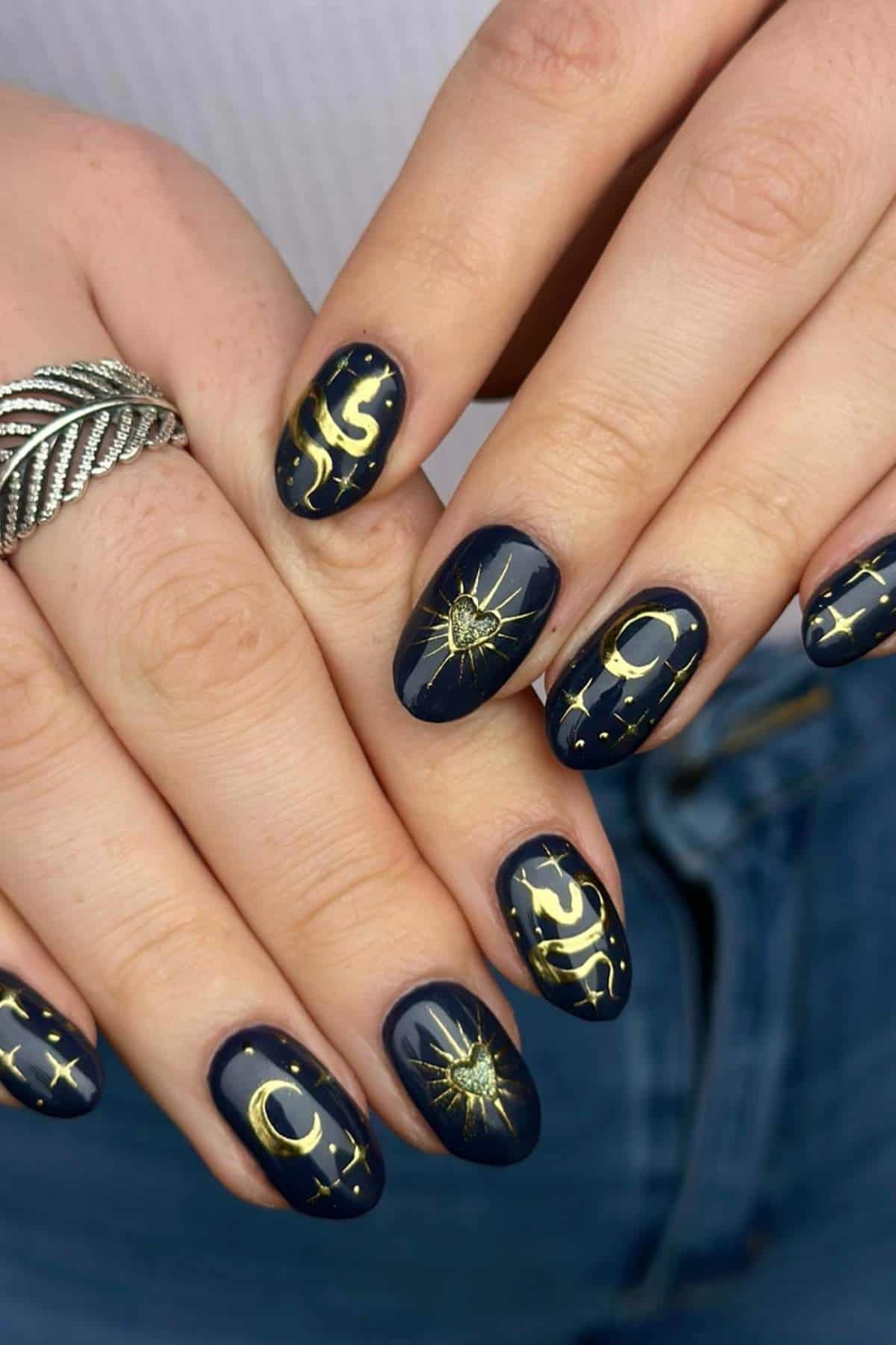 Hands with dark blue nail polish featuring gold celestial designs of moons, stars, and suns. The person is wearing a silver twisted ring and blue jeans.