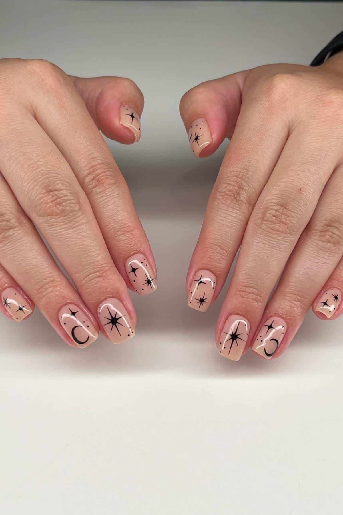 Two hands with nude-colored nail polish featuring black celestial designs, including stars and crescent moons, displayed on a white surface.