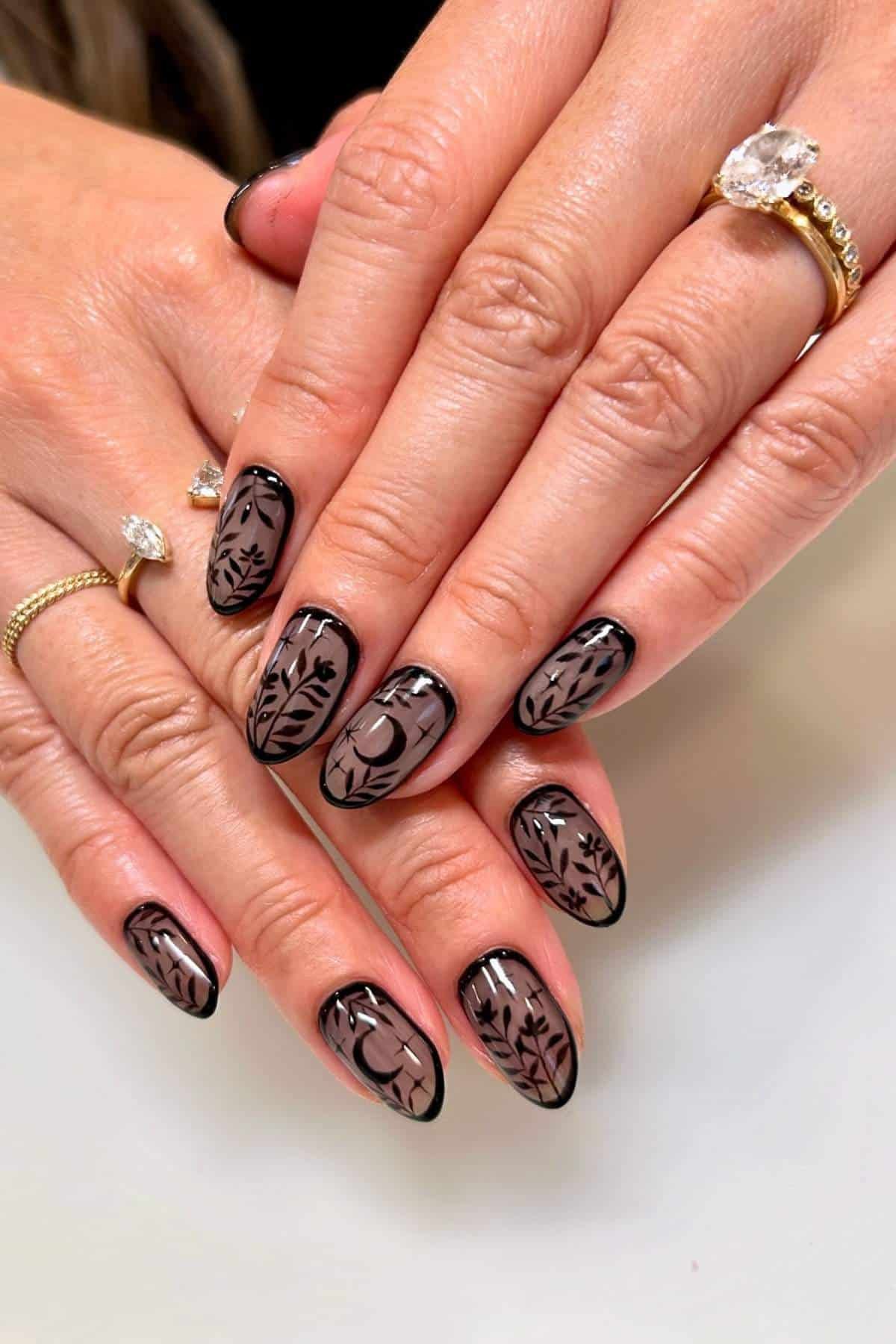 Hands with almond-shaped nails featuring black botanical designs over a nude base. The person wears gold rings, one with a large clear gemstone.