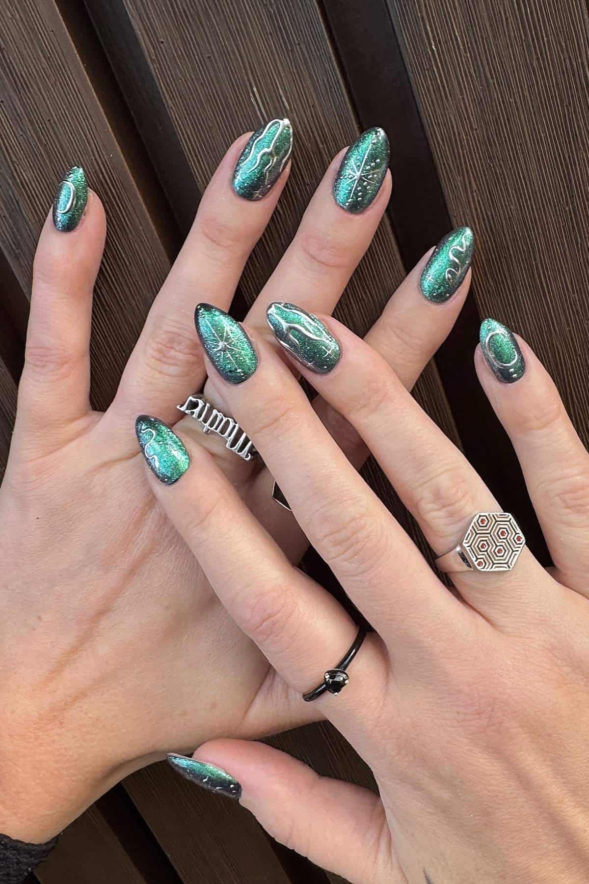 Hands with long, oval-shaped nails painted metallic green with marble designs, wearing three rings, displayed against a brown textured background.