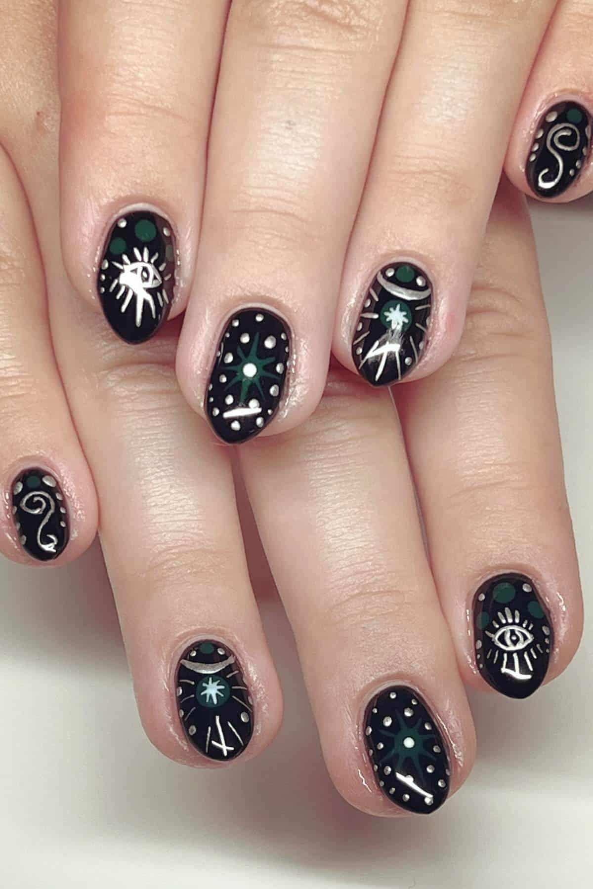 Fingernails painted black with white and green celestial designs, including stars, eyes, and abstract shapes, on both hands resting on a flat surface.