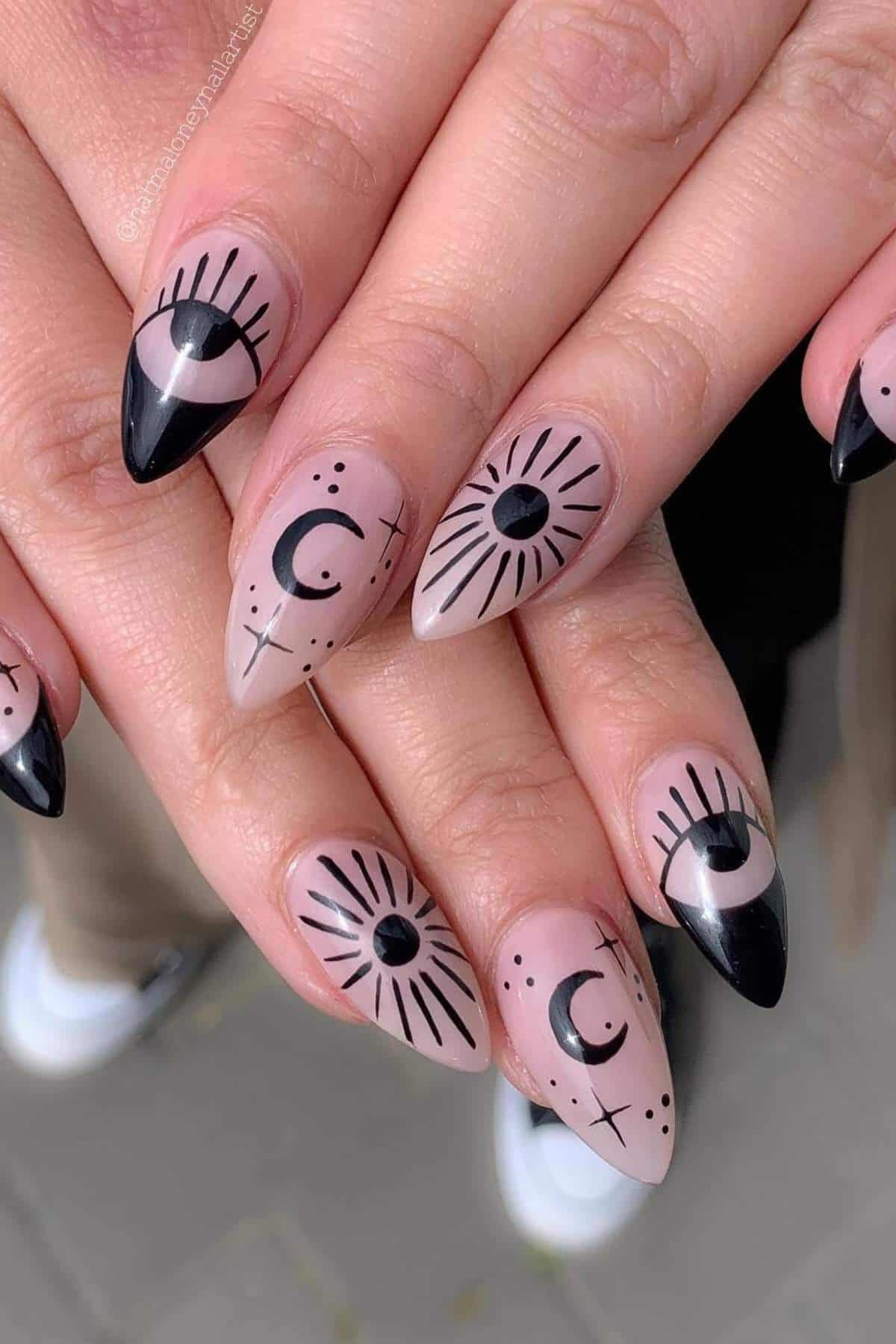Hands with almond-shaped nails painted with black eye, moon, and star designs on a nude base. The tips are black with artistic line details. The background shows part of feet in white shoes.